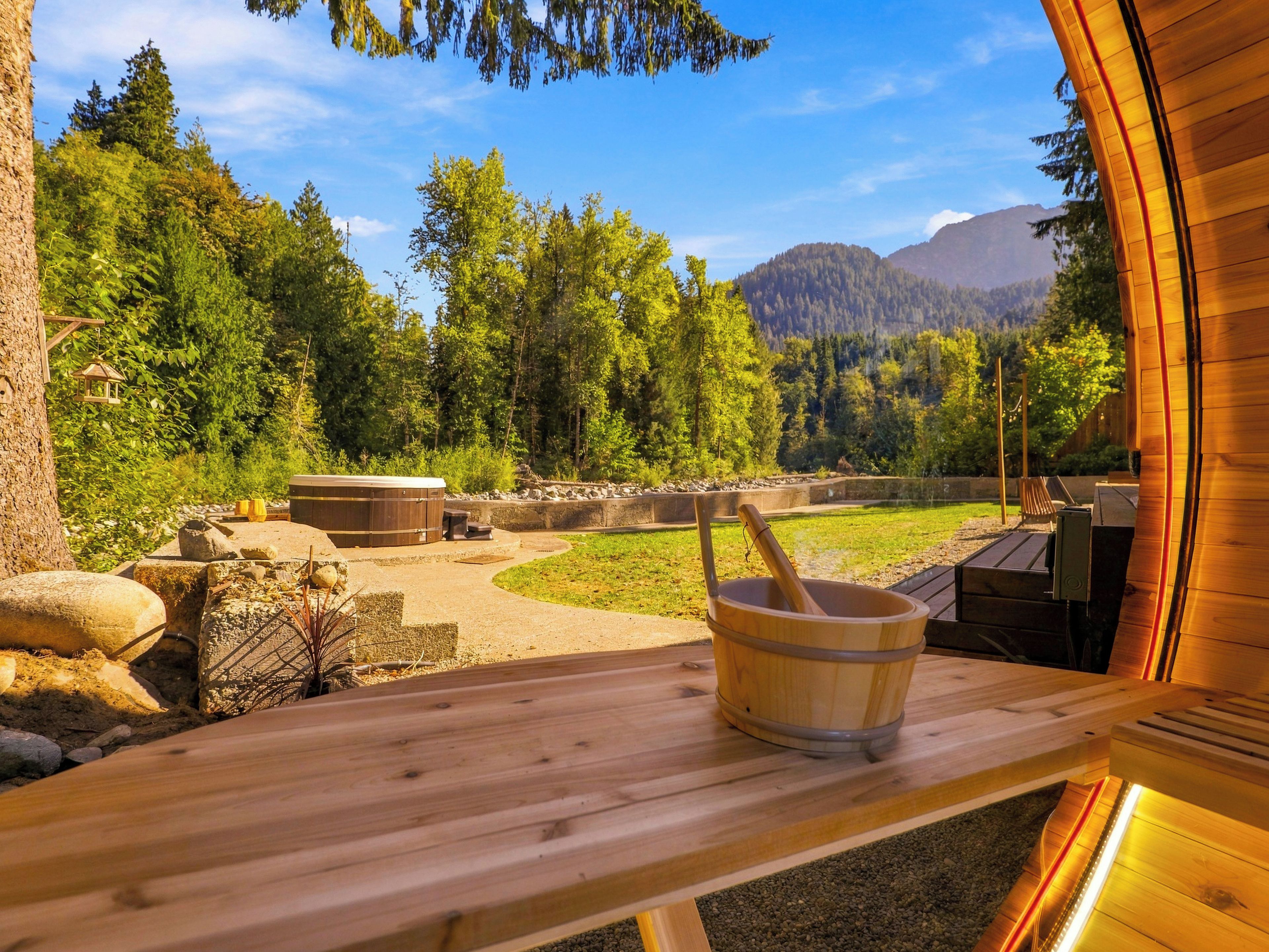 Un banco de sauna de madera con un balde de madera en el centro, rodeado de un paisaje natural con árboles y montañas al fondo.