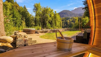 Un banco de sauna de madera con un balde de madera en el centro, rodeado de un paisaje natural con árboles y montañas al fondo.