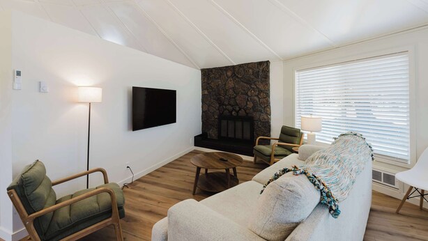 A living room with a flat-screen TV, a stone fireplace, a sofa, a chair, and a window with blinds. - Central Carousel (Bend)