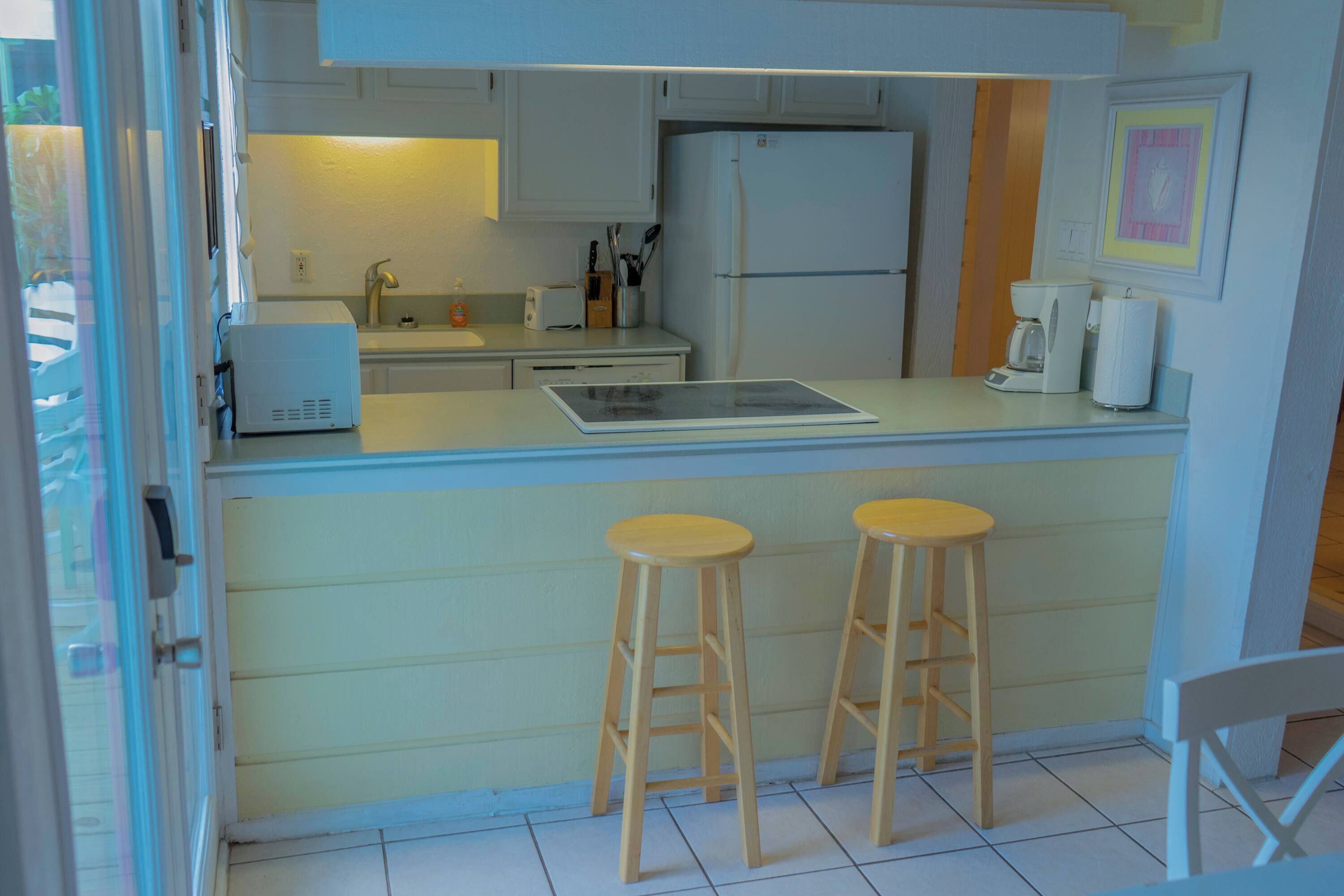 Eine kleine Küche mit weißen Schränken, einem Einbaukühlschrank, einer Kaffeemaschine und zwei Holzhockern.