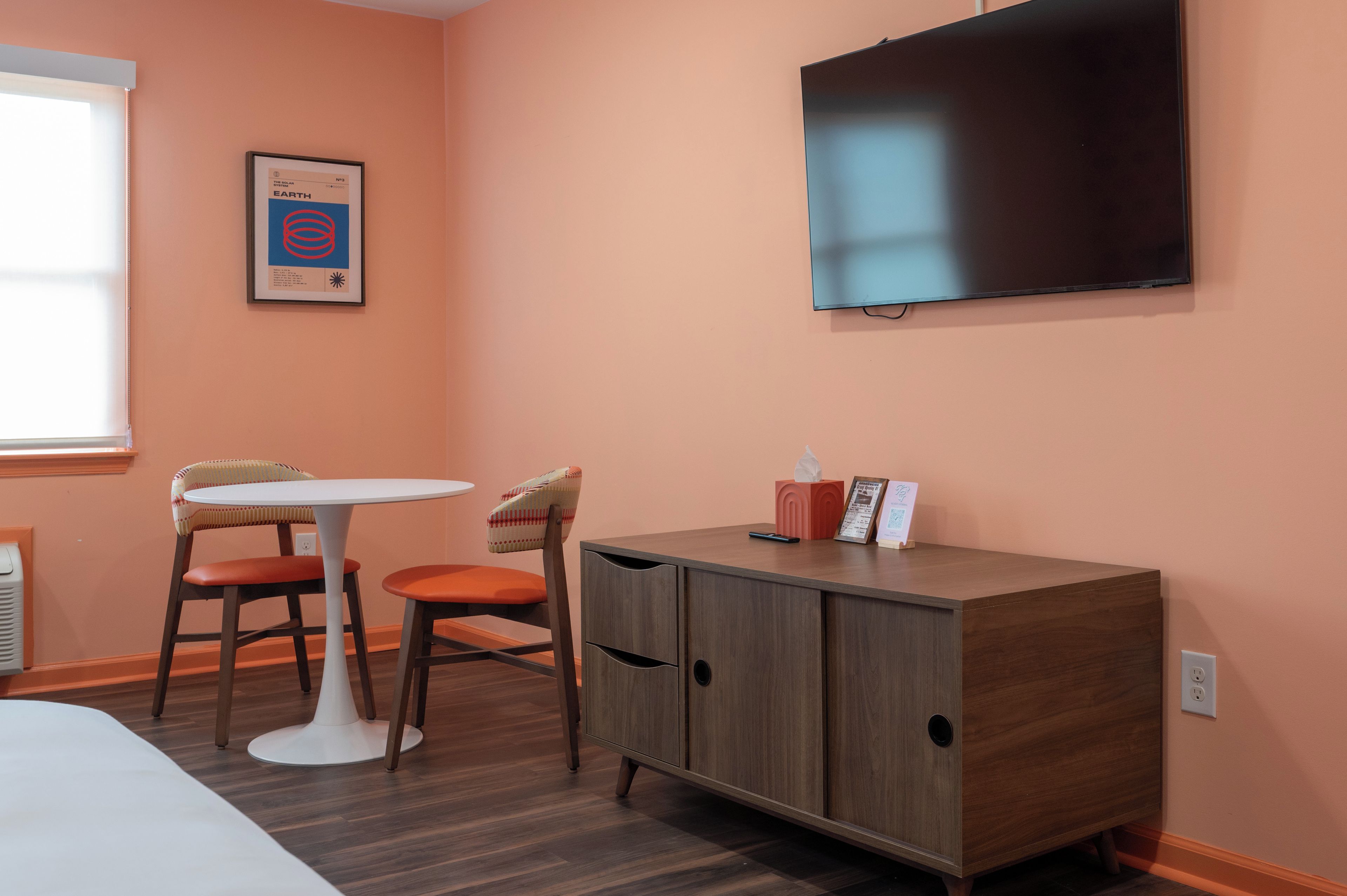 Une chambre avec un lit simple, une petite table avec deux chaises, une armoire, un téléviseur fixé au mur et un tableau encadré accroché au mur.