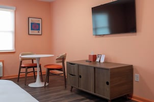 Una habitación con una cama individual, una mesita con dos sillas, un armario, un televisor montado en la pared y un cuadro enmarcado.