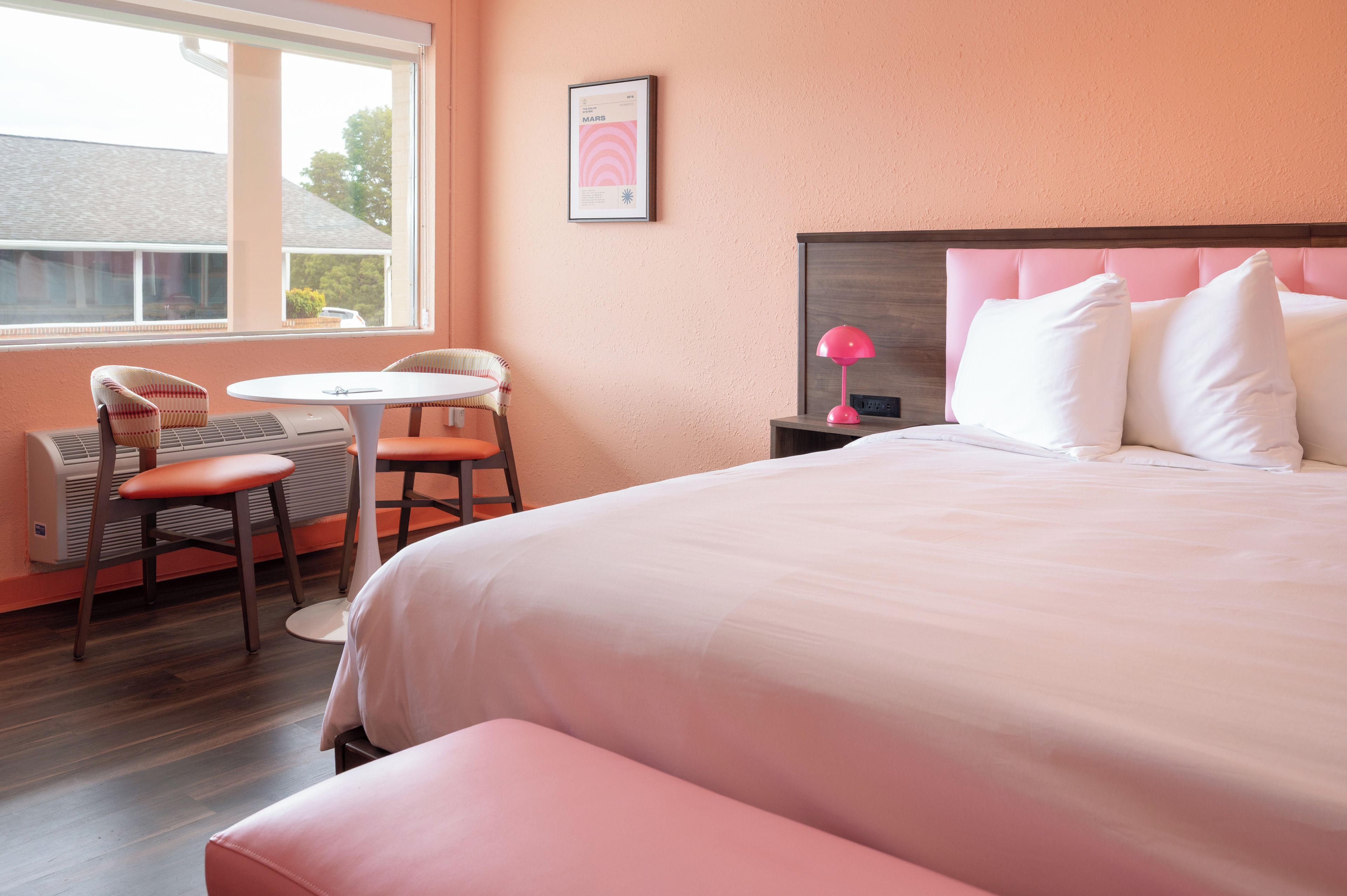 A hotel room with a bed, a desk, two chairs, a window, and a nightstand with a lamp.