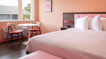 A hotel room with a bed, a desk, two chairs, a window, and a nightstand with a lamp.