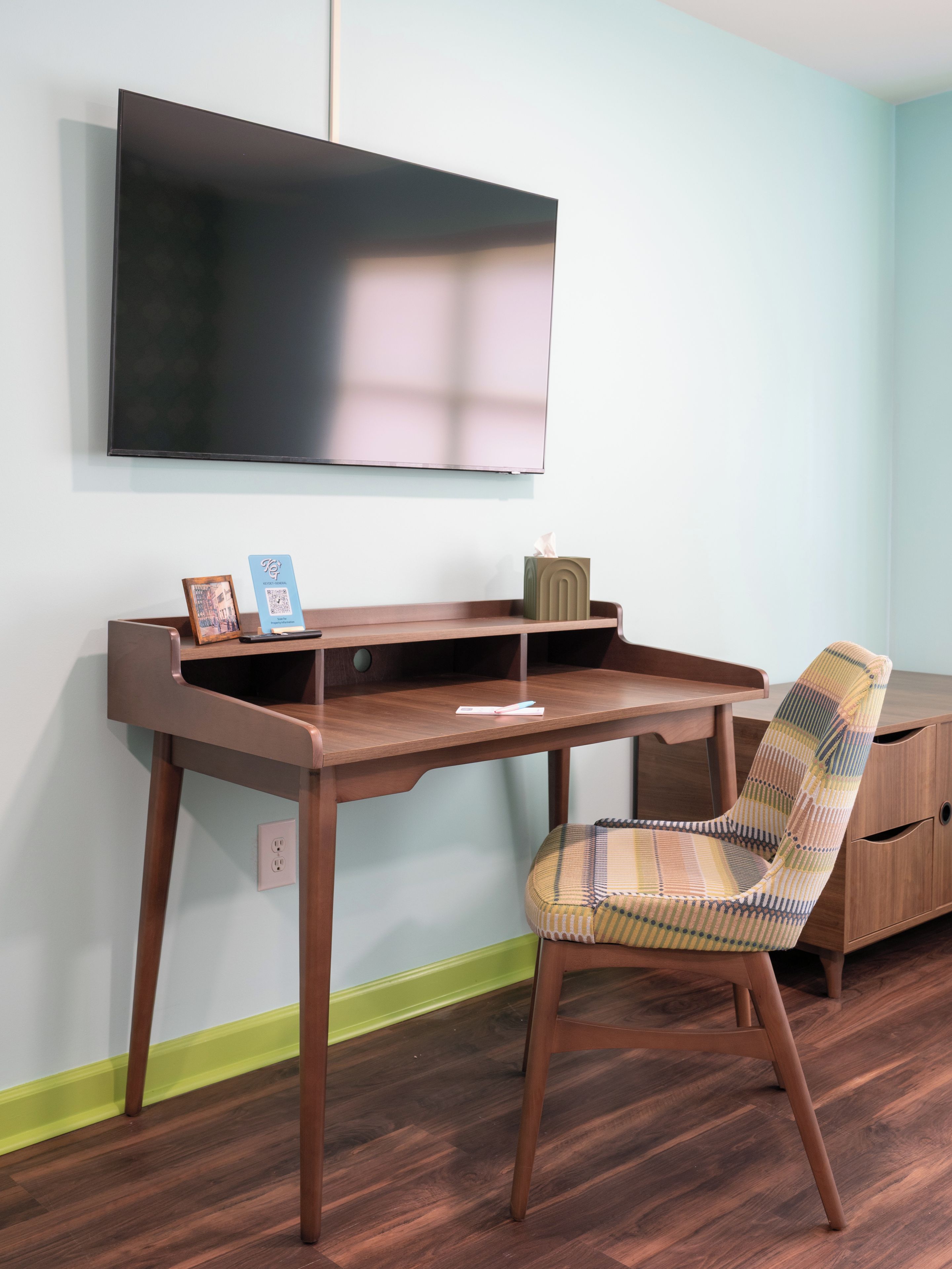 A room with a wooden desk, a chair, a television mounted on the wall, and a small cabinet.