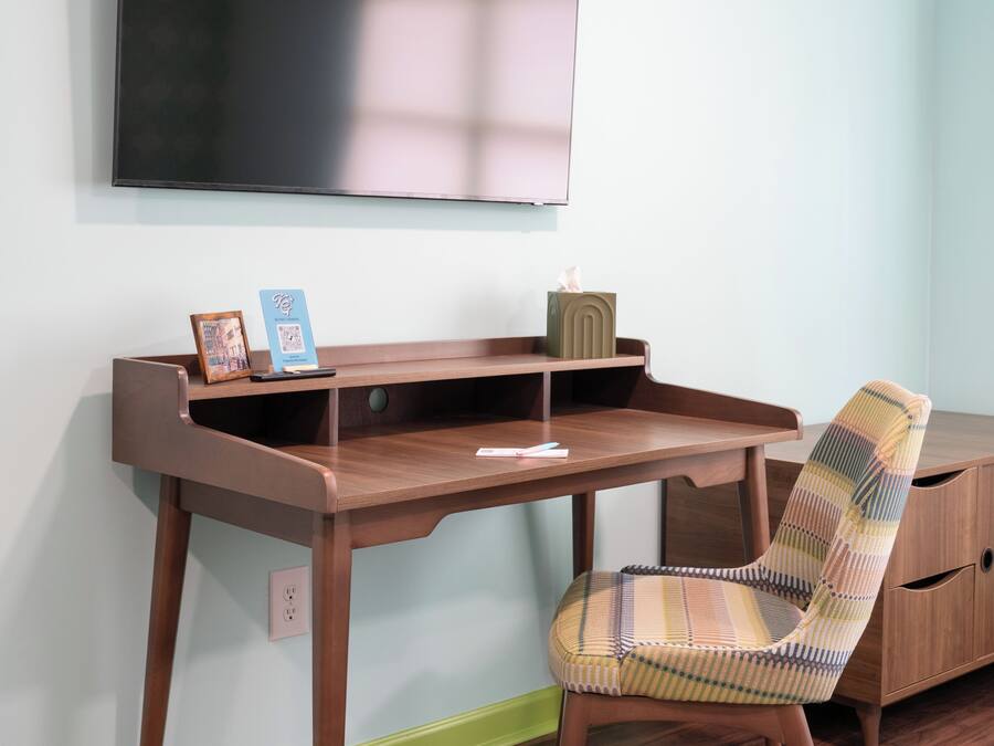 Una habitación con un escritorio de madera, una silla, un televisor montado en la pared y un mueble bajo.