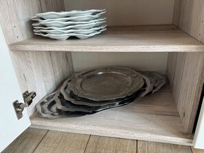 A wooden cabinet with two shelves; the top shelf holds stacked plates, and the bottom shelf contains a stack of black plates. - Maison accessible PMR tout confort ,Jardin,proche de la plage et du centre ville (Saint-Brevin-les-Pins)