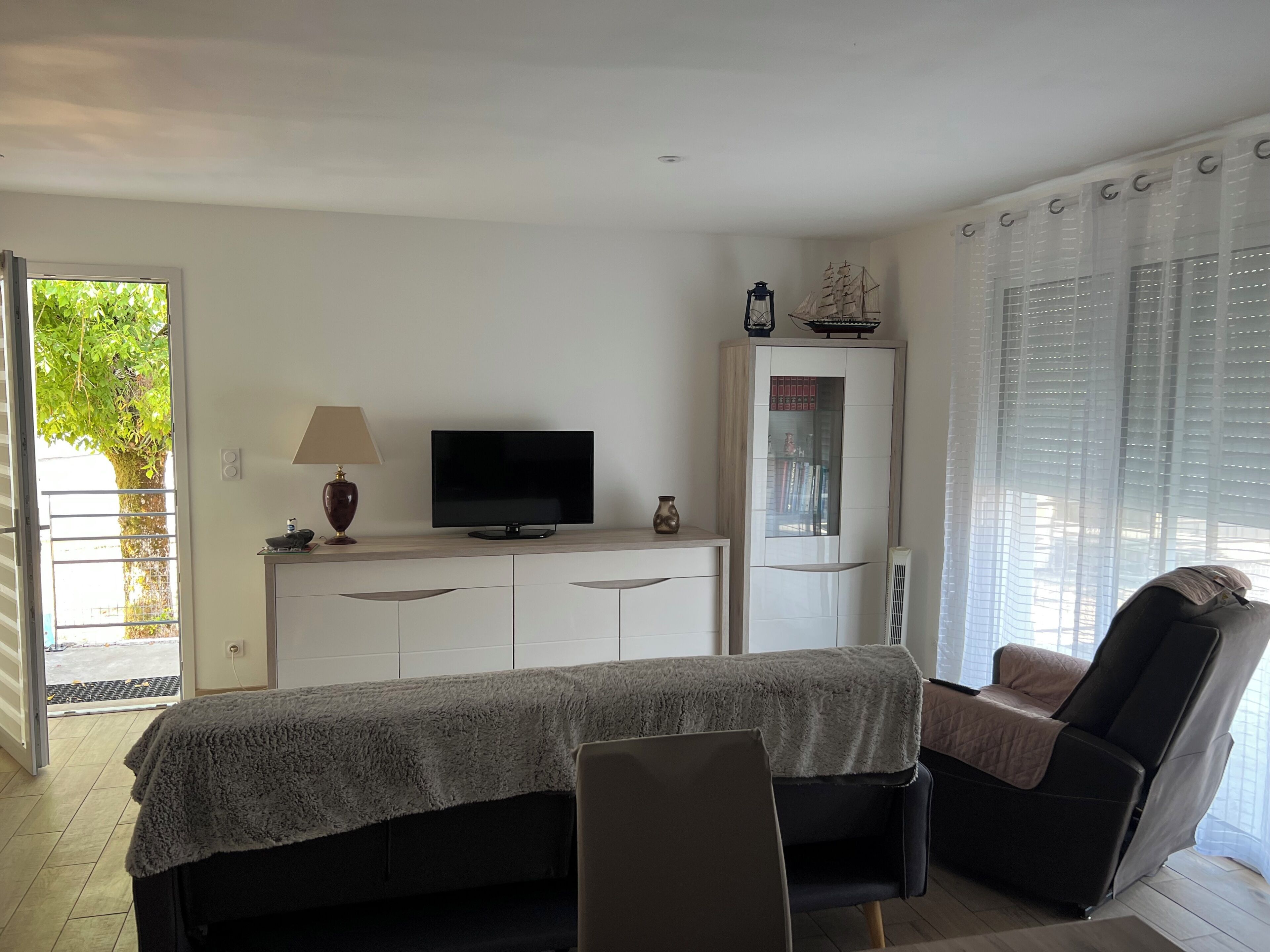 Un salon moderne comprenant un téléviseur à écran plat, un canapé, un fauteuil, un meuble orné d’un objet décoratif et une fenêtre avec des stores.