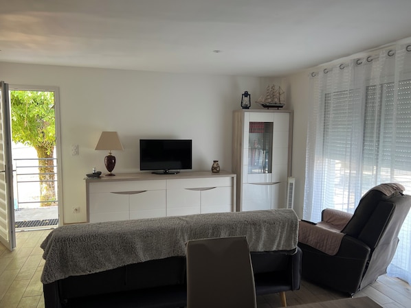 Ein modernes Wohnzimmer mit Fernseher, Sofa, Sessel, Schrank mit Dekoration und Fenster mit Jalousien.