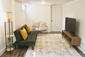 A living room with a black sofa, a wooden TV stand, a patterned rug, and a chair with cushions. - Luma House - Poolside 4-bedroom retreat in Moncton (Moncton)