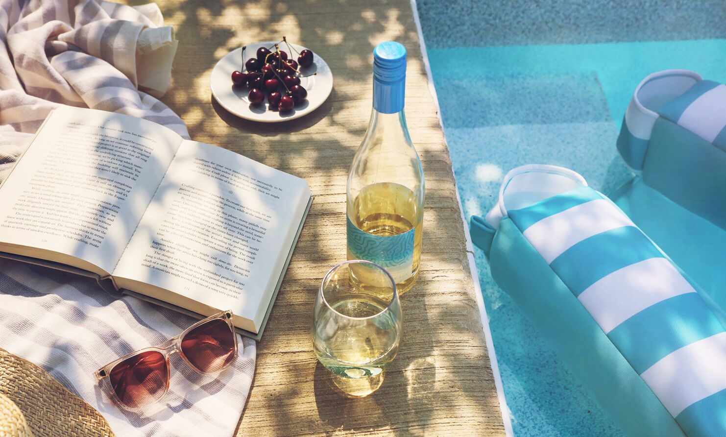 A book, a bottle of wine, a glass of wine, sunglasses, a plate of cherries, and striped cushions by a pool.