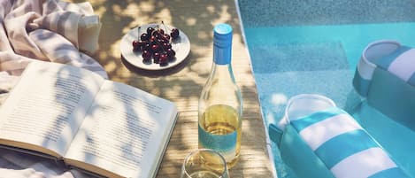 A book, a bottle of wine, a glass of wine, sunglasses, a plate of cherries, and striped cushions by a pool.