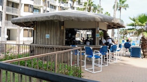 A beachside bar with a thatched roof, blue chairs, and a view of a hotel in the background. - Direct Beachfront 3BR - Access to 3 Pools & 3 Hot Tubs (South Padre Island)