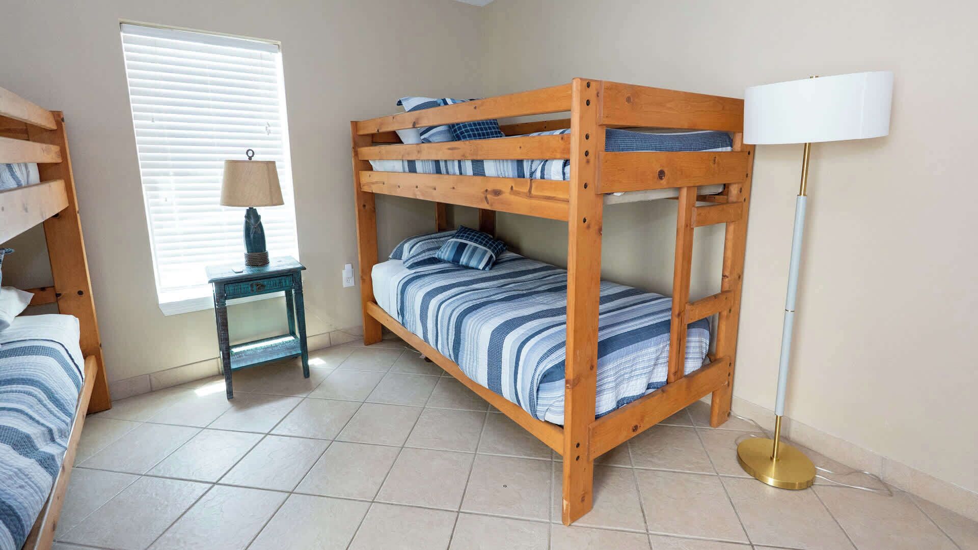 A bunk bed room with a lamp, a small table, and a window with blinds.