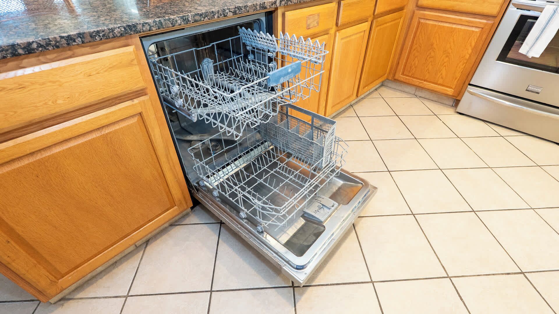 A kitchen with wooden cabinets, a stainless steel dishwasher, and a tiled floor.