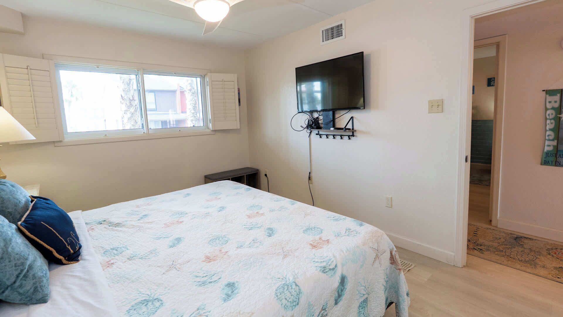 Un dormitorio con cama, televisor montado en la pared, ventana con contraventanas y puerta que da a otra habitación.