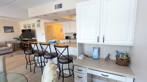 A modern kitchen with white cabinets, a dining area with bar stools, and a living room with a sofa and TV. - 2BR Condo - 2 Pools - Jacuzzi - Steps to Beach SPI (South Padre Island)