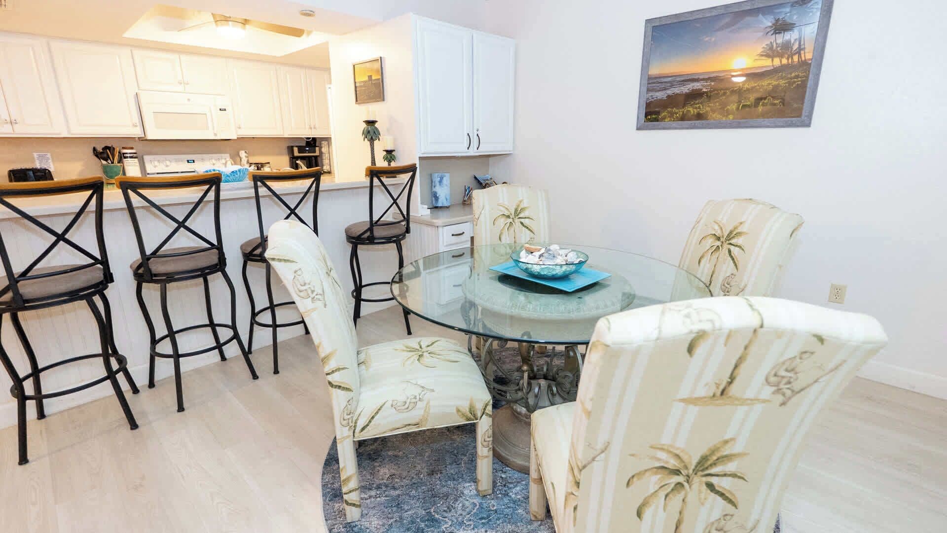 A dining area with a glass table surrounded by chairs, a kitchen in the background, and a sunset painting on the wall.