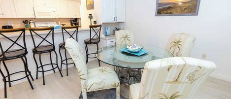 A dining area with a glass table surrounded by chairs, a kitchen in the background, and a sunset painting on the wall.