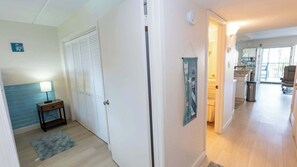 A hallway with a closed door, a small table with a lamp, a blue rug, and a poster on the wall. - 2BR Condo - 2 Pools - Jacuzzi - Steps to Beach SPI (South Padre Island)