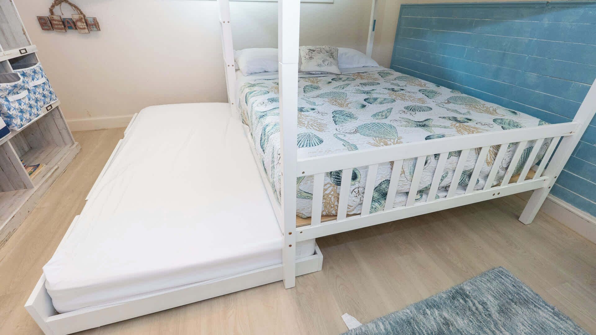Un lit superposé avec un matelas sur le lit du bas, un berceau blanc posé sur le sol, un mur bleu et une étagère en bois avec des livres.