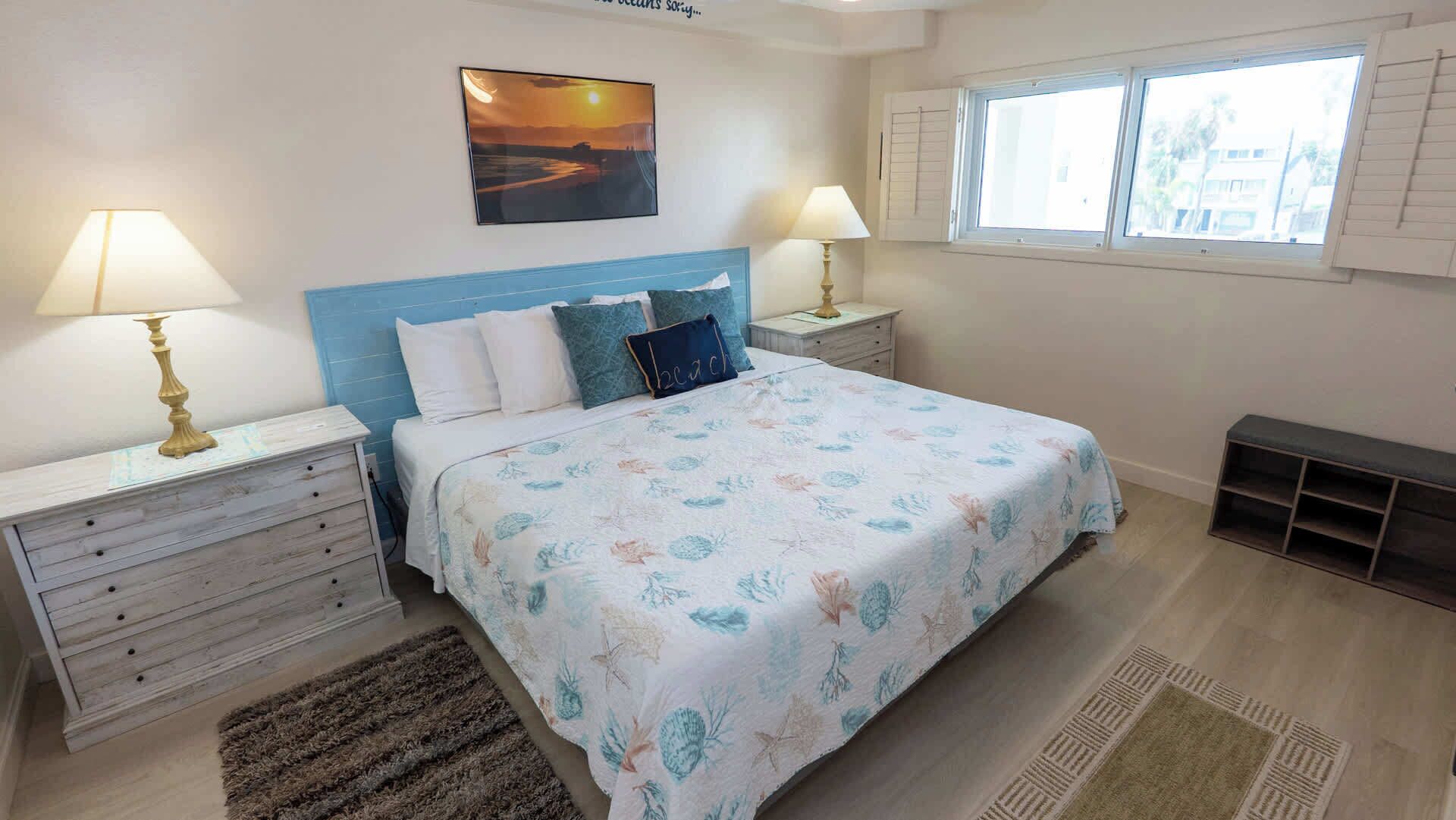 A bedroom with a bed, two bedside tables, a nightstand, a window with shutters, and a painting on the wall.
