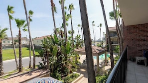 A balcony with a view of palm trees, a pool, and a brick building. - 2BR Condo - 2 Pools - Jacuzzi - Steps to Beach SPI (South Padre Island)