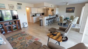 A living room with a TV, sofa, and coffee table, a kitchen with white cabinets, and a dining area with chairs. - 2BR Condo - 2 Pools - Jacuzzi - Steps to Beach SPI (South Padre Island)