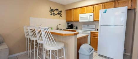 Cocina compacta con refrigerador, microondas y estufa. Hay tres sillas blancas alrededor de una pequeña isla.