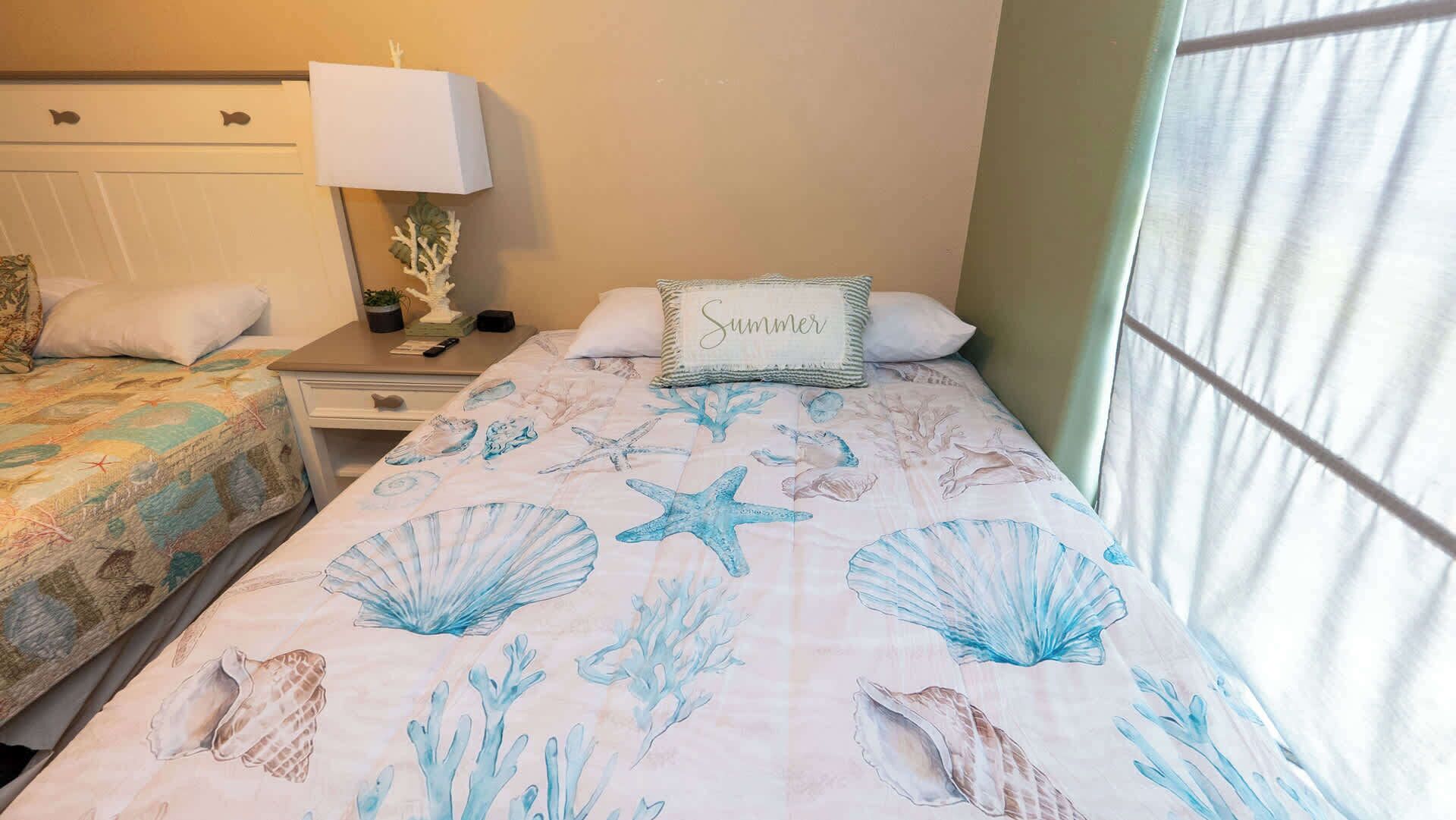 A bedroom with two beds, each with a different sea-themed bedspread, a bedside table with a lamp, and a nightstand with a small plant.