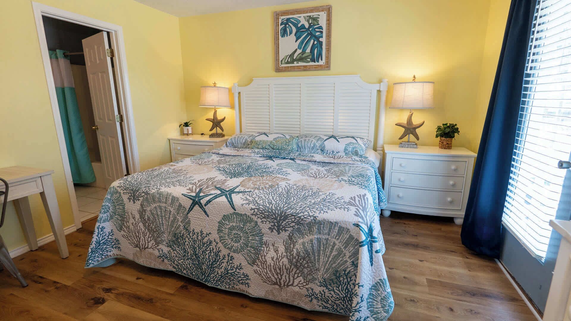 Um quarto com cama, duas mesas de cabeceira, cômoda, porta e janela com cortinas.