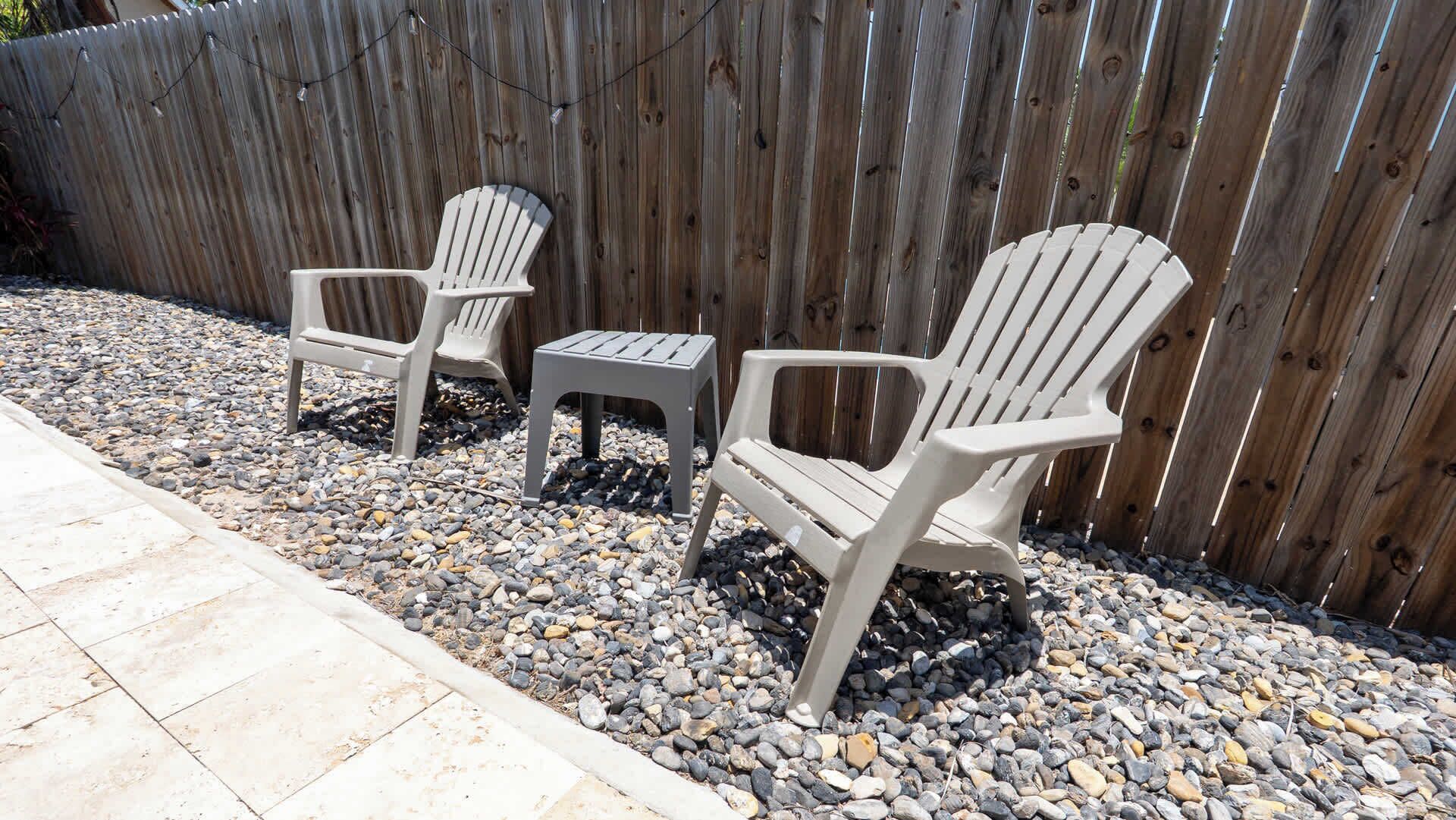 Two Adirondack chairs and a small table set on a gravel surface, with a wooden fence in the background.