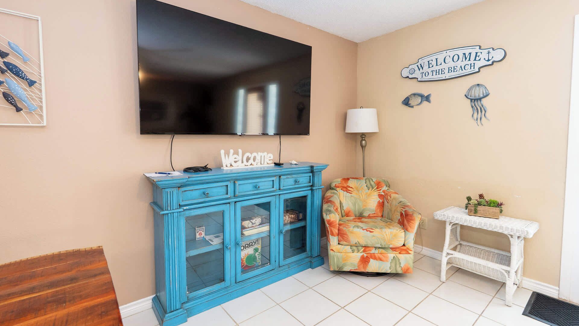Una sala de estar con televisor, un mueble azul, un sillón estampado, una mesita con plantas y un cartel de bienvenida en la pared.