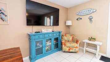 Uma sala de estar com televisão, um armário azul, uma poltrona florida, uma mesinha com plantas e um aviso de boas-vindas na parede.