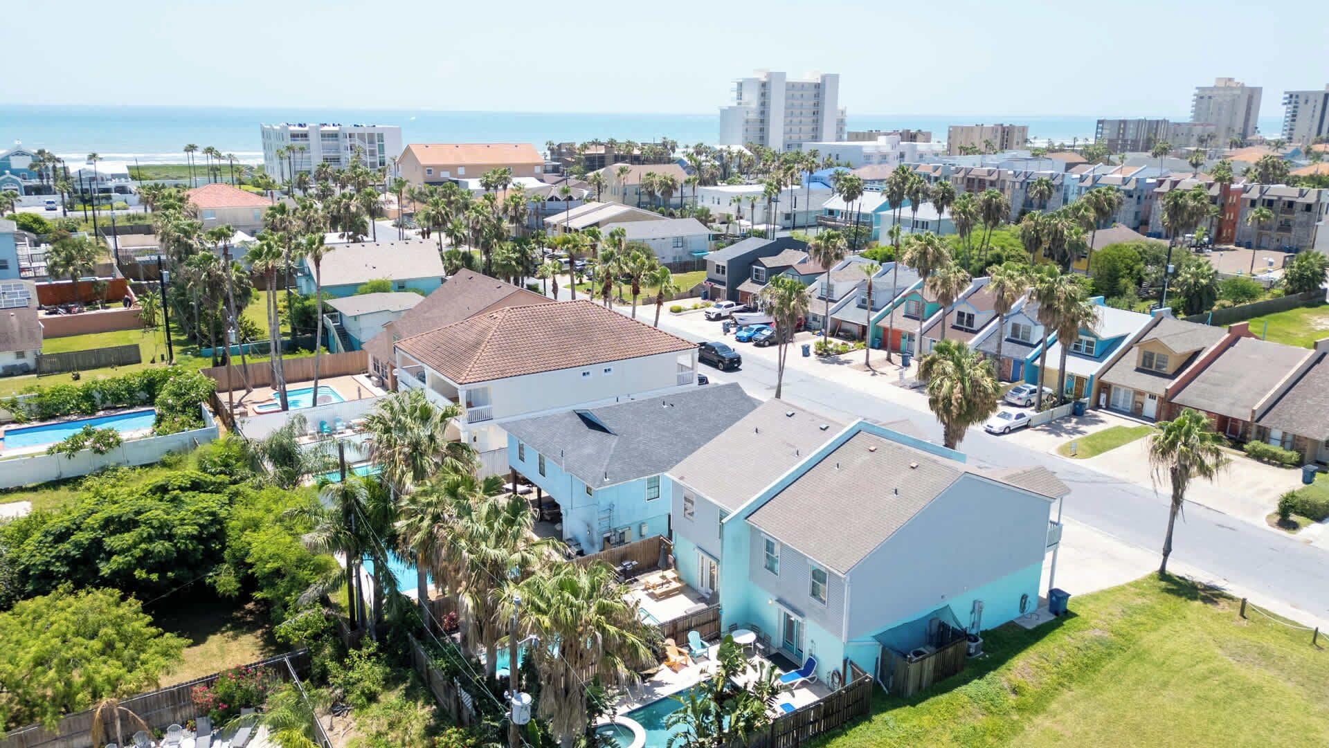 A residential area with a large house featuring a swimming pool, surrounded by palm trees and a view of the ocean.