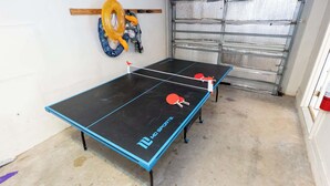 Une table de ping-pong avec deux raquettes, un anneau gonflable jaune et une serviette bleue et blanche à motifs, accrochée au mur.