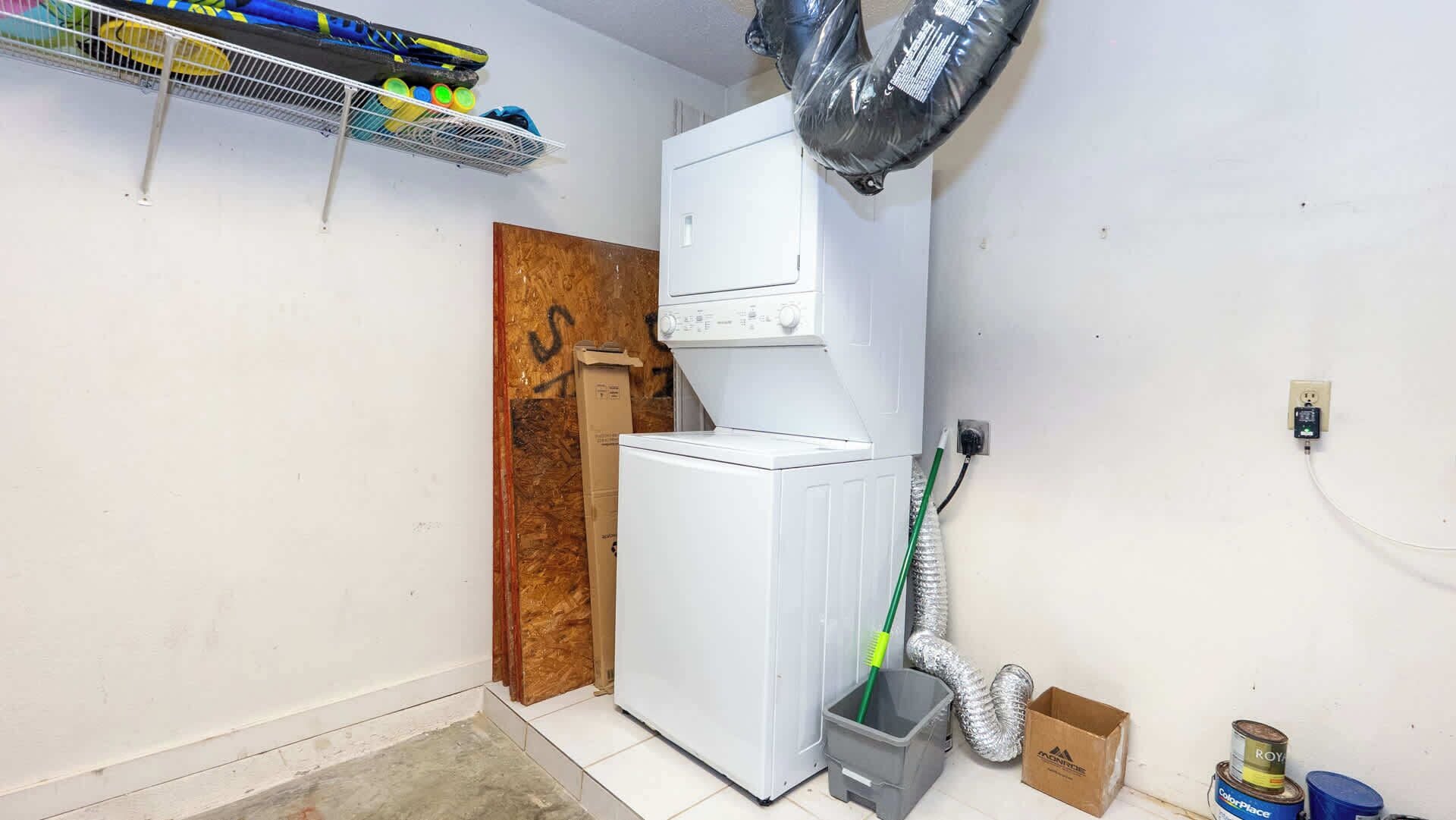 Een wasserette met een witte wasmachine en droger, een houten deur, een metalen plank met schoonmaakmiddelen en een ventilatieslang.