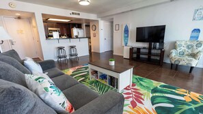 A living room with a sofa, a coffee table, a TV, a chair, and a kitchen area in the background. - Beachfront 2BR 11th Floor Views, Saida Royale SPI (South Padre Island)