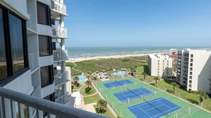 Een hoog hotel met balkons met uitzicht op een strand, een zwembad en tennisbanen.