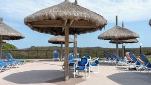 A beachside lounge area with thatched umbrellas and blue lounge chairs. - Beachfront 2BR 11th Floor Views, Saida Royale SPI (South Padre Island)