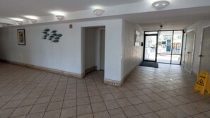 Um corredor amplo com piso ladrilhado, uma porta que dá para o exterior e um aviso no chão.