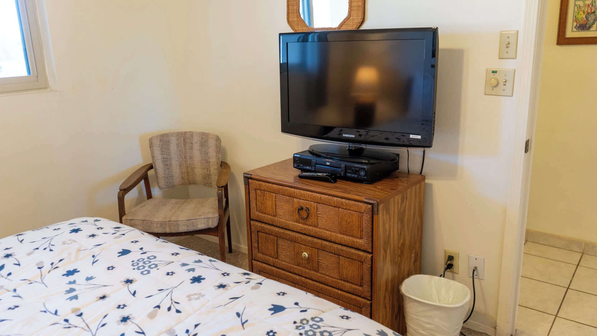 A bedroom with a bed, a chair, a wooden dresser with a television and audio equipment, and a white trash bin.