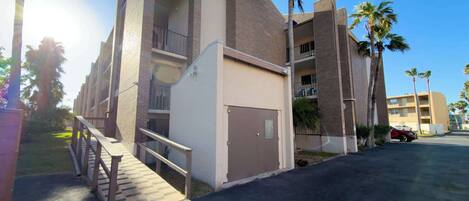 A multi-story building with a central staircase, balconies, and a parking area with palm trees.