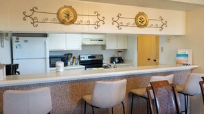 Eine Küche mit weißen Schränken, einem Kühlschrank, einer Mikrowelle, einem Herd und einer Kaffeemaschine. Es gibt drei Barhocker und eine Wand mit dekorativen Tellern.