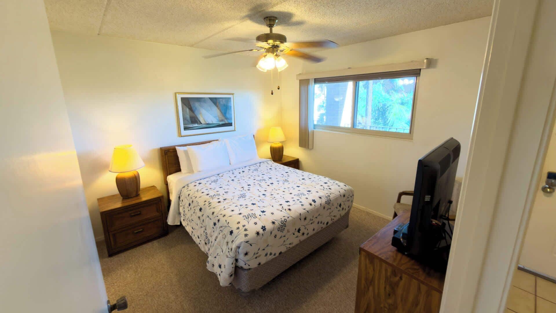 A bedroom with a bed, two bedside lamps, a ceiling fan, a window, and a television on a wooden stand.