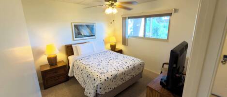 A bedroom with a bed, two bedside lamps, a ceiling fan, a window, and a television on a wooden stand.