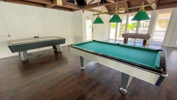 Une table de billard recouverte d’un drap vert, une table de billard et un porte-queue dans une pièce au plancher en bois et dotée d’un éclairage suspendu vert.
