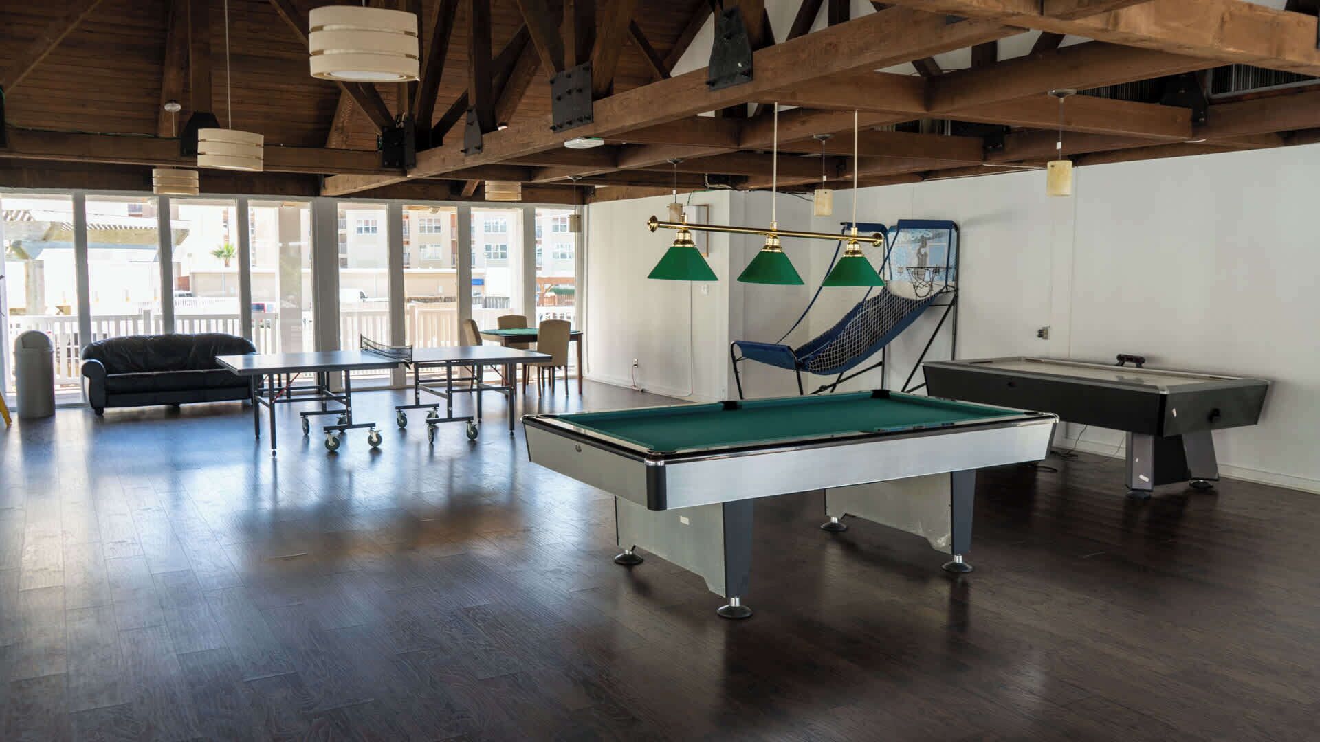 Une table de billard recouverte d’un drap vert, un queue de billard et un hamac bleu dans une pièce dont le plafond est orné de poutres en bois et qui possède de grandes fenêtres.