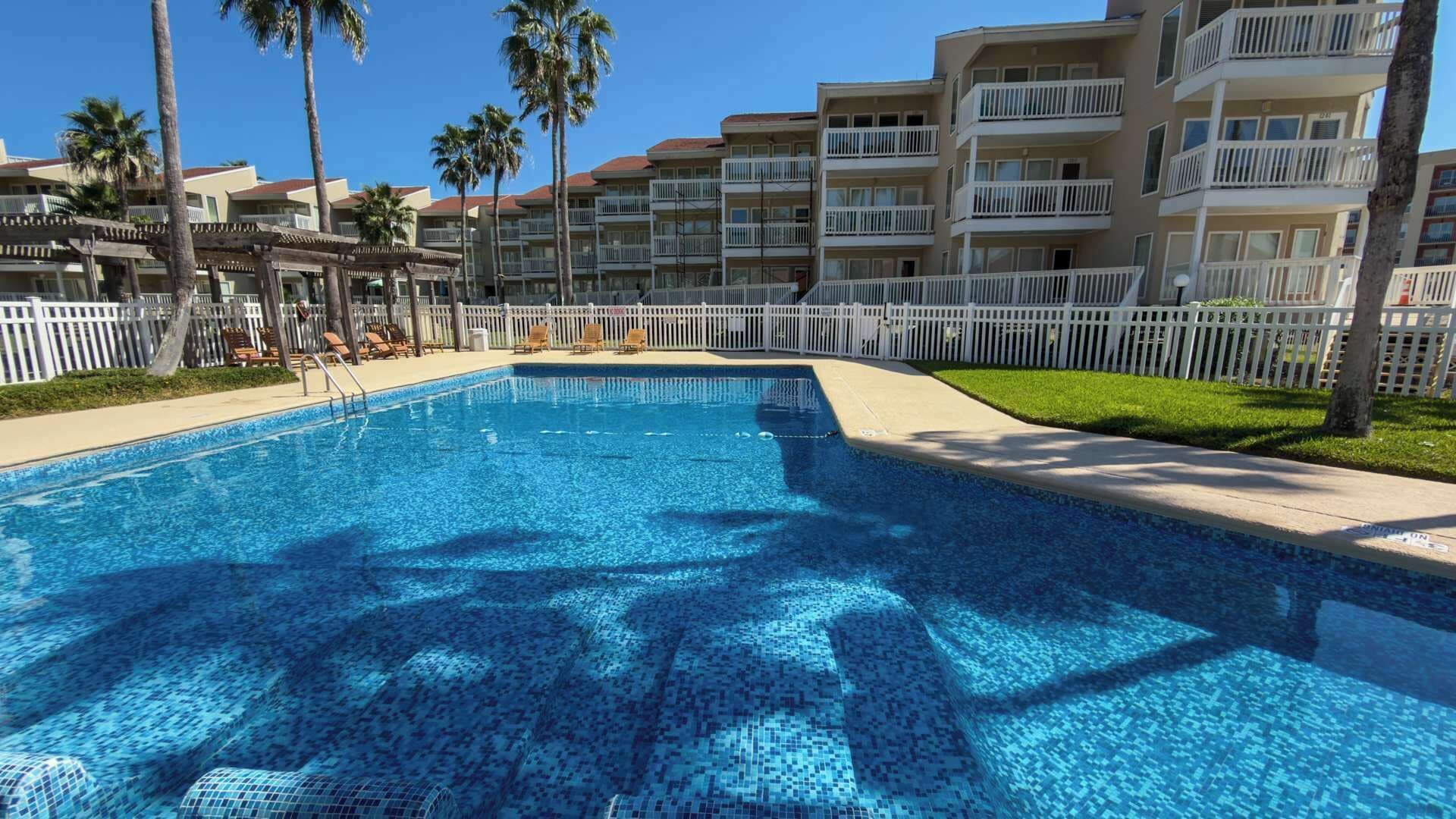 A swimming pool with a tiled deck, surrounded by a white fence, palm trees, and multi-story buildings with balconies.