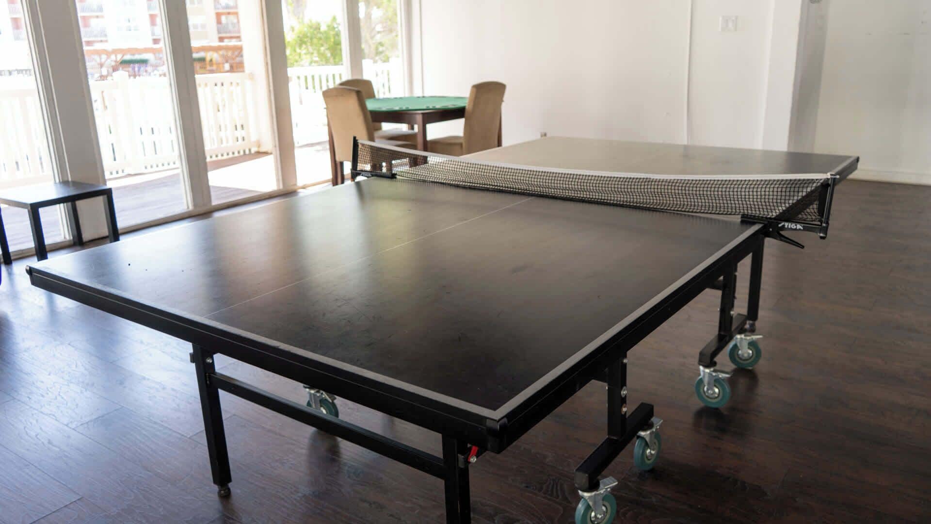 Taula de ping-pong sobre rodes en una habitació amb terra de fusta i vista a un balcó amb mobles de jardí.